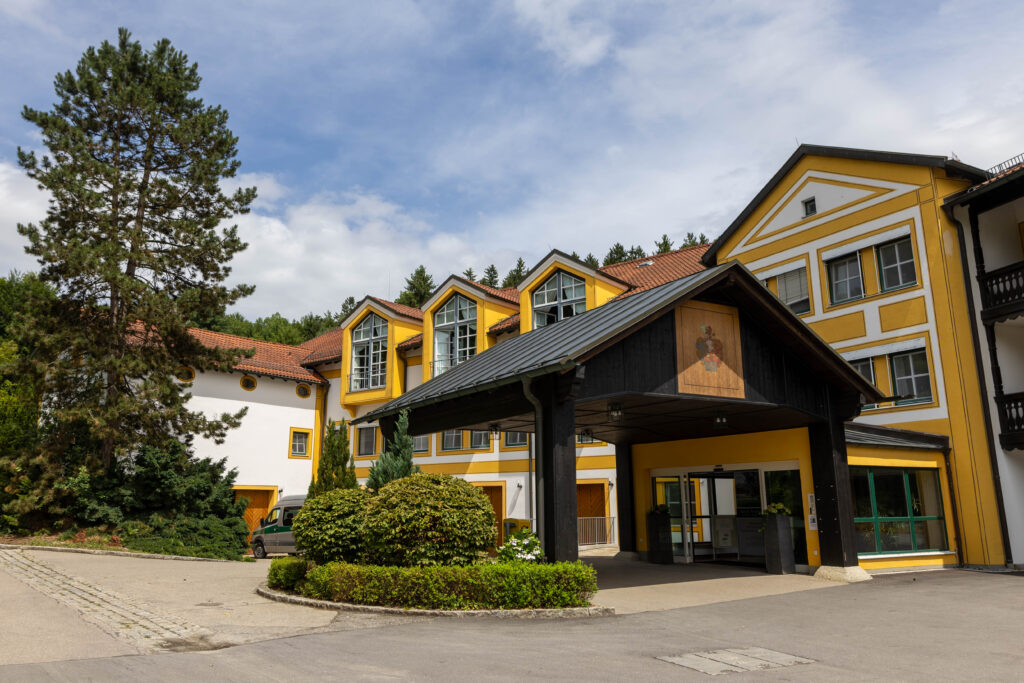 Foto des Haupteingangs der Klinik Wartenberg. Man sieht den vorgelagerten, überdachten Bereich vor der Tür. Vor diesem befinden sich grüne Hecken. Im Hintergrund erkennt man das Hauptgebäude der Klinik, das gelb-weiß gestrichen ist. Links im Bild sieht man einen großen Nadelbaum. Im Vordergrund sieht man die asphaltierte Zufahrt zum Eingangsbereich. Der Himmel ist blau und leicht bewölkt.
