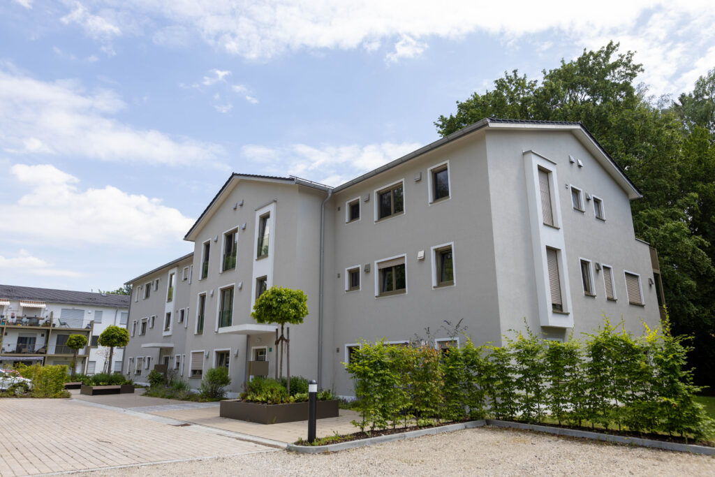 Foto der klinikeigenen Betriebswohnungen am Bründlplatz in Wartenberg. Ein graues Wohnhaus steht auf dem begrünten Gelände. Davor der gepflasterte Parkplatz. Im Hintergrund Bäume.