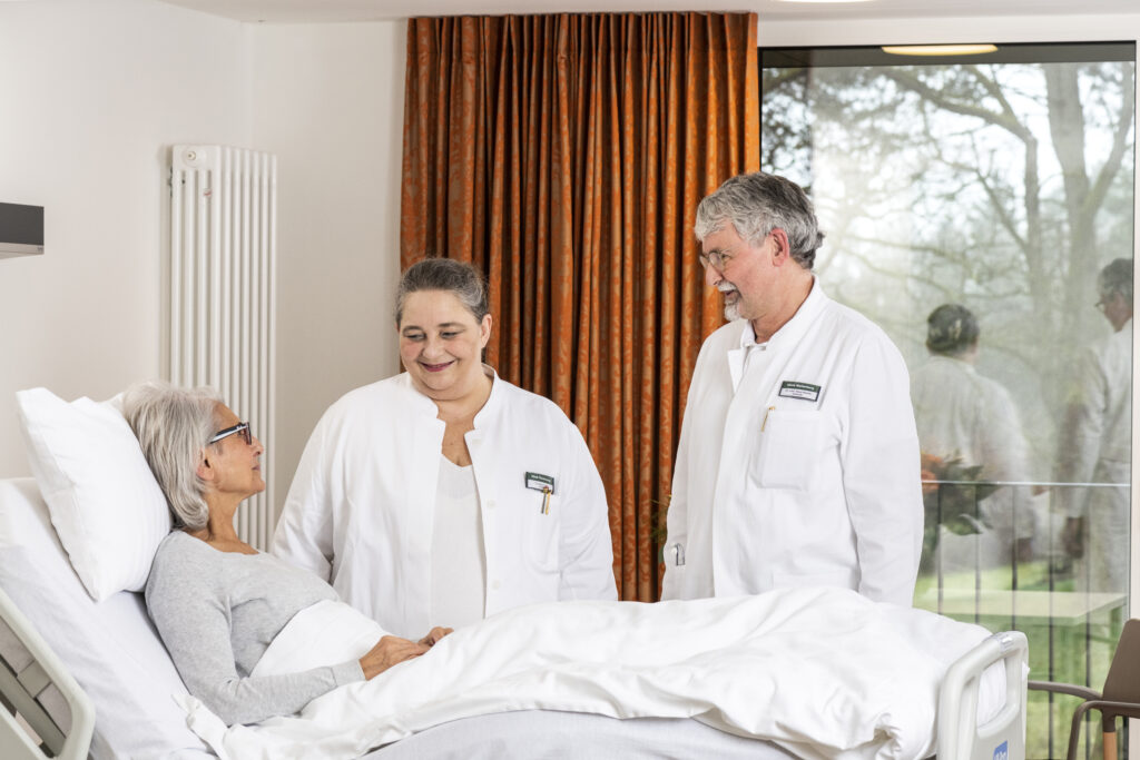 Auf dem Foto sieht man die beiden Chefärzte der Klinik Wartenberg, Frau Doktor Rupp und Herrn Doktor Becher, neben dem Patientenbett, in dem eine Patientin liegt. Die beiden Ärzte schauen die Patientin an und lächeln. Im Hintergrund sieht man das Fenster im Patientenzimmer, durch welches man in den Innenhof schauen kann.