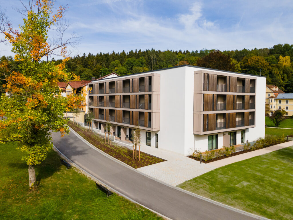 Auf dem Foto sieht man den Neubau der Klinik Wartenberg. Links im Bild steht ein Baum mit Blättern in grün-gelb-orangenen Farben, es scheint Herbst zu sein. Im Hintergrund erkennt man die weiteren Gebäudeteile der Klinik. Dahinter befindet sich der Klinikwald.