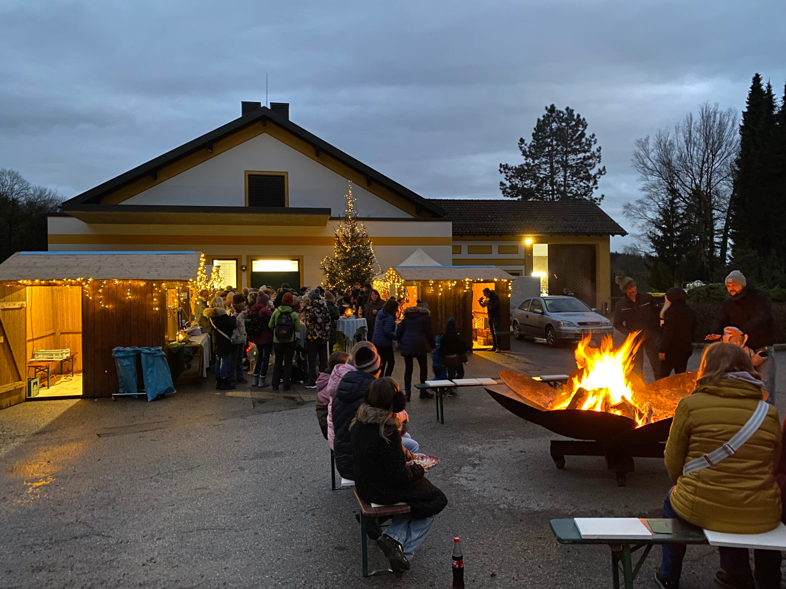 Foto der Weihnachtsfeier 2024 auf dem Platz hinter der Küche auf dem Klinikgelände der Klinik Wartenberg. Die Weihnachtsfeier ist wie ein kleiner Weihnachtsmarkt aufgebaut. Man sieht mehrere Buden, in denen Speisen, Getränke und Selbstgemachtes verkauft wird. Viele Kolleginnen und Kollegen haben sich auf dem Platz versammelt. Vorne rechts im Bild sieht man eine Feuerschale, an der sich Mitarbeitende aufwärmen. Die Weihnachtsfeier findet traditionell jedes Jahr in der Vorweihnachtszeit für die Mitarbeitenden der Klinik Wartenberg statt.