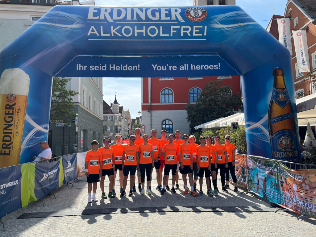 Foto des Teams der Klinik Wartenberg vom Stadtlauf Erding 2024. Auf dem Bild stehen die Läuferinnen und Läufer in ihren leuchtend-orangenen Trikots unter dem Startbogen vom Stadtlauf Erding.