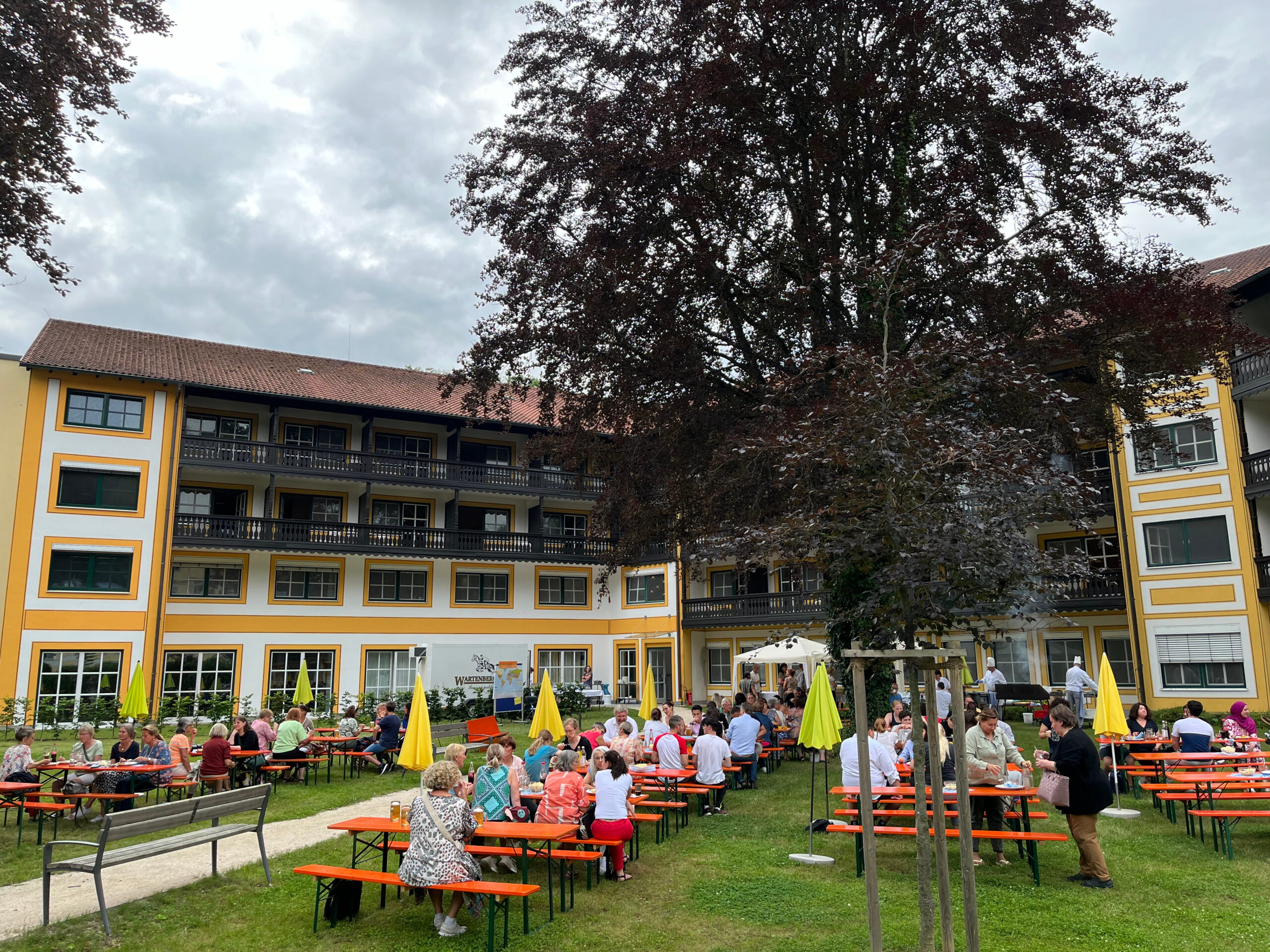 Foto vom Sommerfest 2024 der Klinik Wartenberg im Innenhof der Klinik Wartenberg. Kolleginnen und Kollegen sitzen auf Bierzeltgarnituren, essen und trinken. Im Hintergrund sieht man die Mitarbeitenden von Küche und Service, die grillen und sich um die gastronomische Versorgung kümmern. Das Sommerfest findet jedes Jahr für die Mitarbeitenden der Klinik Wartenberg statt.