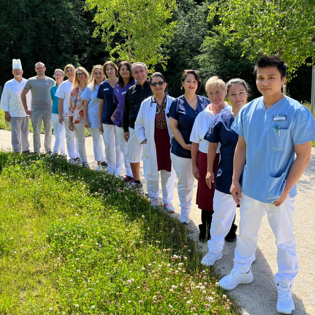 Foto der Klinik Familie auf dem Klinikgelände. Eine Gruppe von Mitarbeitenden verschiedener Berufsgruppen steht hintereinander aufgereiht auf einem Weg auf dem Klinikgelände. Hintergrund des Fotos ist es, die Vielfalt der Klinik Wartenberg zu repräsentieren. Links vorne im Bild sieht man da grüne Gras im Innenhof, im Hintergrund erkennt man die Bäume des Klinikwaldes.