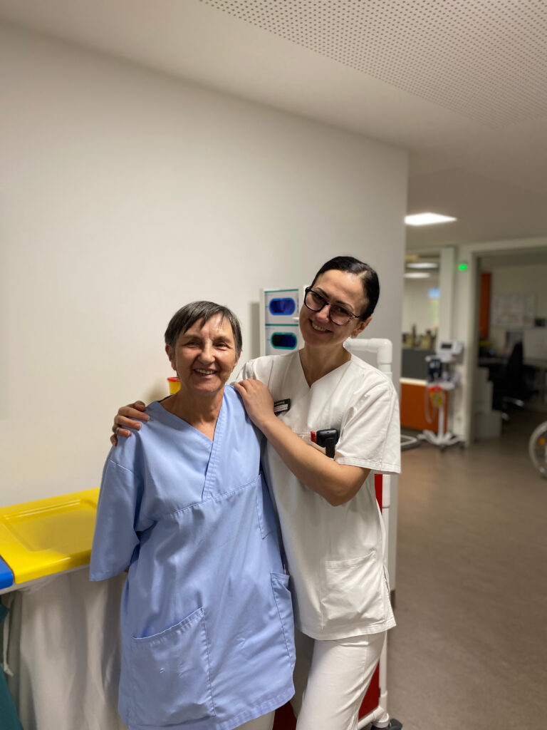 Foto der beiden Integrationsbeauftragten Roza Angelovski und Dijana Smickovic, die sich gegenseitig im Arm halten. Sie stehen auf einer Akutstation im Flur, tragen Arbeitskleidung und lächeln in die Kamera. Im Hintergrund steht ein Pflegewagen und rechts hinten sieht man das den Stationsstützpunkt.