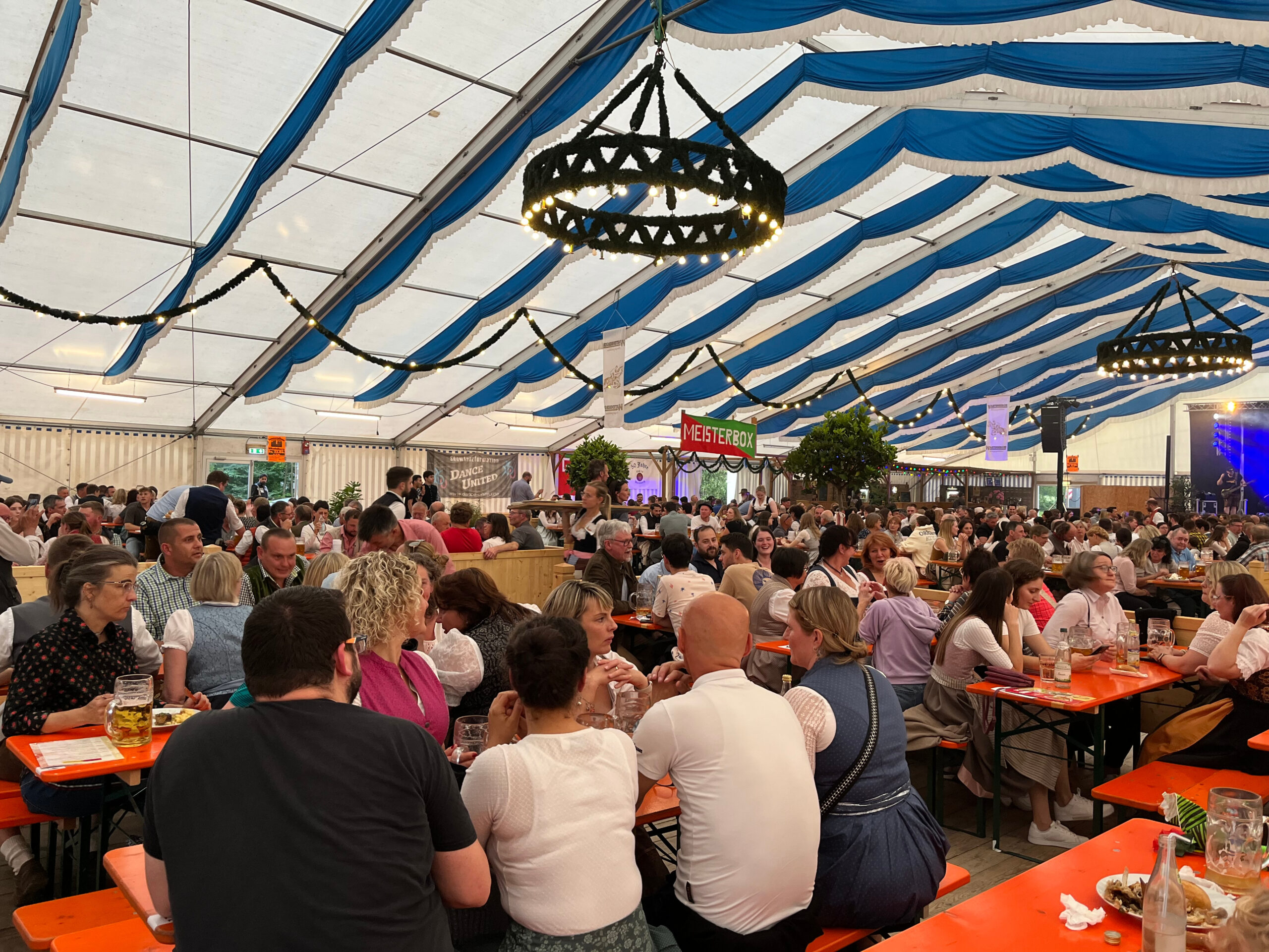 Foto vom Volksfest Wartenberg 2024. Kolleginnen und Kollegen verbringen einen unterhaltsamen Abend auf dem "Tag der Betriebe" beim Volksfest Wartenberg. Viele tragen Trachten. Die Mitarbeitenden sitzen auf Bierzeltgarnituren im Festzelt. Das Zelt ist gut gefüllt mit vielen Betrieben aus Wartenberg. Das Zeltdach ist blau-weiß und es hängen beleuchtete Kränze an der Decke. Im Hintergrund kann man die Bühne erkennen.