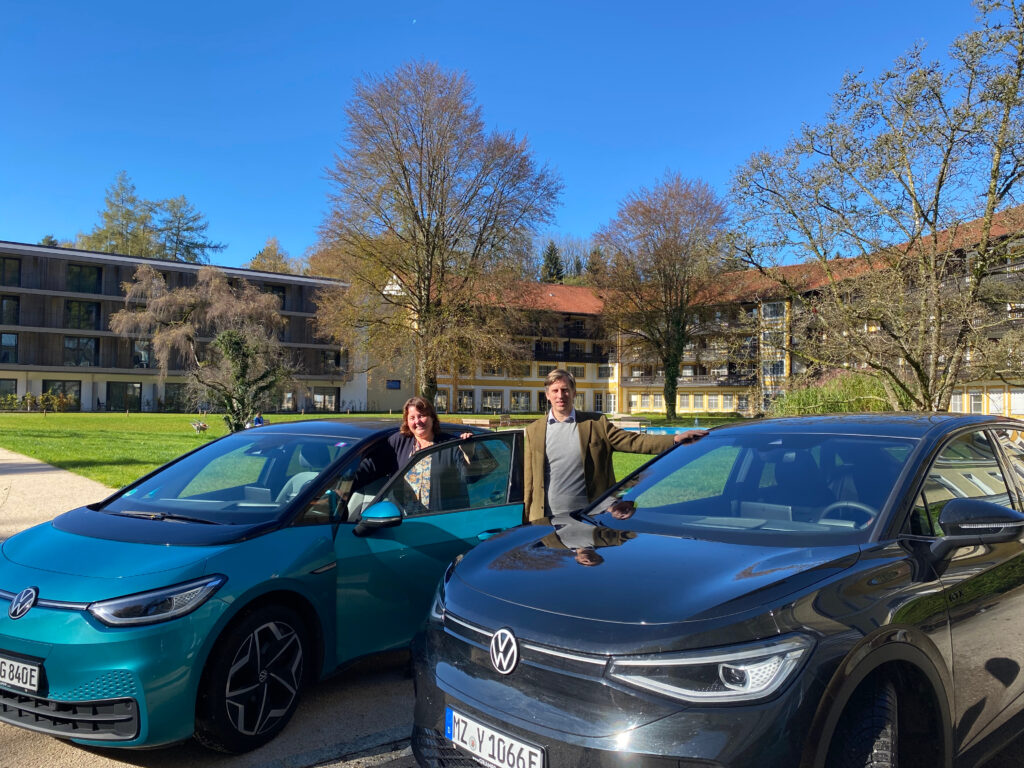 Foto von zwei Elektroautos von VW im Innenhof der Klinik Wartenberg. Die Personalleiterin und der Geschäftsführer stehhen mittig zwischen den Autos. Im Hintergrund sieht man das Klinikgebäude und den strahlend blauen Himmel. Dieses Foto wurde für das Benefit des E-Auto-Leasings aufgenommen, welches Mitarbeitenden der Klinik Wartenberg geboten wird.