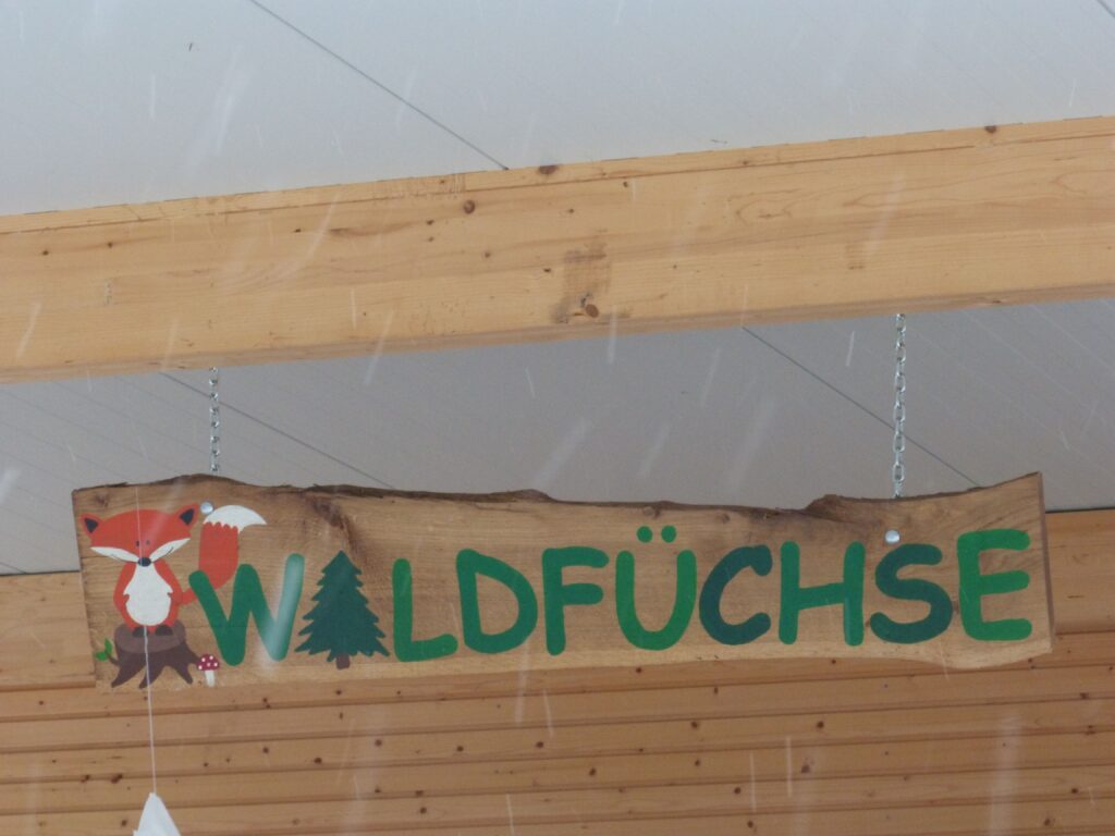 Foto des Schildes "Waldfüchse", welches im Waldkindergarten am Gebäude auf dem Klinikgelände hängt. Das Schild ist aus einem Holzbrett gemacht und auch das Gebäude ist hölzern.